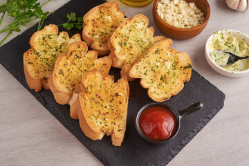 Homemade tasty bread with garlic, cheese and herbs on kitchen table. Baguette with herb butter and Rosemary, parsley and garlic with raw materials over black stone floor, top view