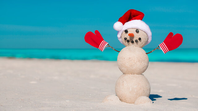 Snowman on the beach. Sandy Christmas snowman in red Santa hat and mittens or gloves. Smiley Snowman at sunny beach. Holiday concept for Happy New Year post cards. Florida Winter. Ocean or Sea water.