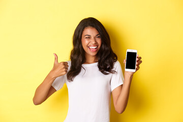 Image of beautiful african-american girl, winking and smiling, showing smartphone screen and...