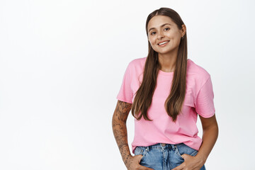 Young stylish woman with tattoos and piercing, smiling and looking confident, standing against white background