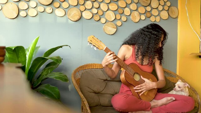 Mujer morena de cabello rizado tocando guitarra con su mascota en una banca en un hostal