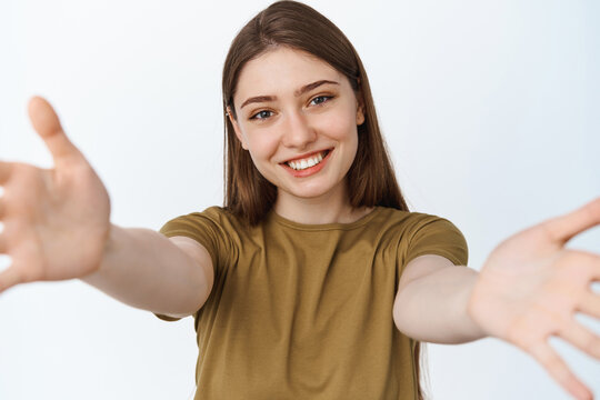 Close up portrait of happy girl hugging you, stretching out hands towards camera and smiling, reaching arms to hold something, standing over white background