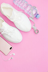 A fitness concept with a water bottle, sneakers, a phone with headphones, a purple fitness elastic band, a pink roller on a pink background. Copy space.