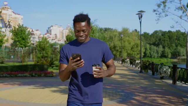 Smiling Afro-American man walks street uses mobile phone holding cup with coffee or tea in hand. Happy African adult male ethnic black wears blue t-shirt looks around and navigates in smartphone