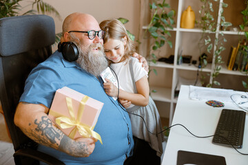 Happy man with gift box embraces blonde little girl holding greeting card in home office