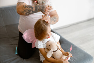 Father with tattoos makes ponytail to little girl wearing fairy suit in room