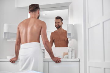 Happy bearded man with bare chest wrapped with towel looks in mirror in modern barthroom
