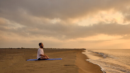 Man practices yoga meditation sitting in padmasana during sunrise. Man sits in lotus position at beach opposite rising sun and its reflection in sea. Portrait of man meditating at beach