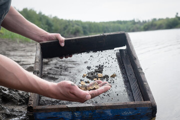 Treasure hunter is picking a golden ore from dirty metal grid on the river water background....