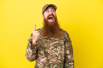 Military Redhead man over isolated on yellow background pointing up and surprised