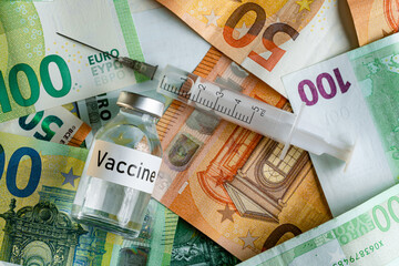 Vaccine price or cost concept - glass vial with silver cap on pile of euro bank notes, syringe near, closeup detail from above