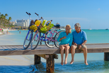 Happy senior couple exercising with bicycles on the beach on a sunny day, sitting on a pier