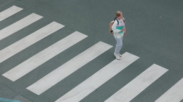 Upper view of smiling trendy pupil in white sweatshirt with workbook, backpack and headphones crossing crosswalk and going to school outdoors in the city.