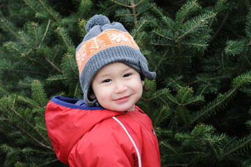 Closeup of adorable happy Asian-American toddler boy smiling at camera outdoors wearing hat and parka with pine tree in background. © Lisa Kim