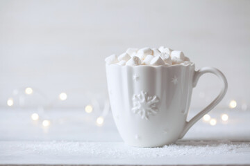 Obraz na płótnie Canvas Christmas card with a mug with a hot drink and marshmallows on a wooden table and a white background, a light garland and space for congratulations text. 