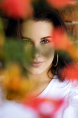 Portrait of a brunette woman hiding behind flowers. A beautiful girl looks seriously huddled against a brick wall and hiding behind flowers. Vertical photo of an attractive woman