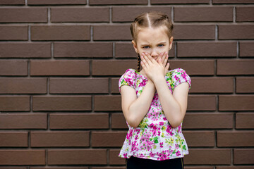 The little girl covers her mouth with her palms against the background of a brick wall. the...