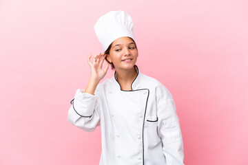 Little caucasian chef girl isolated on pink background listening to something by putting hand on the ear