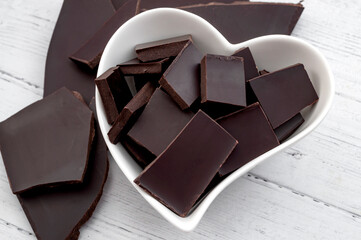 Good source of dietary fiber and cardiovascular health foods concept with dark chocolate pieces in heart shaped ceramic bowl isolated on white wood