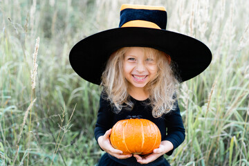 little blonde girl in a black suit and witch hat, Halloween with a pumpkin in her hands