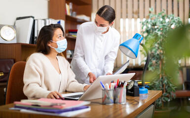 Woman assistant in face mask standing with documents near her female boss in office
