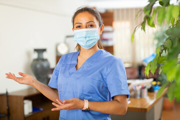Polite female health worker wearing blue uniform and medical face mask welcoming to clinic, making inviting gesture with her hands.