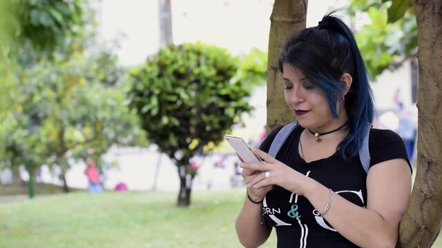 Beautiful serious young woman. blue haired college girl looking at her social media in a park, next to a tree. girl looking at her cell phone in a park. Technology, communication and education concept