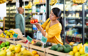 Fototapeta premium Interested Latina choosing ripe red tomatoes while shopping in vegetable department of grocery store