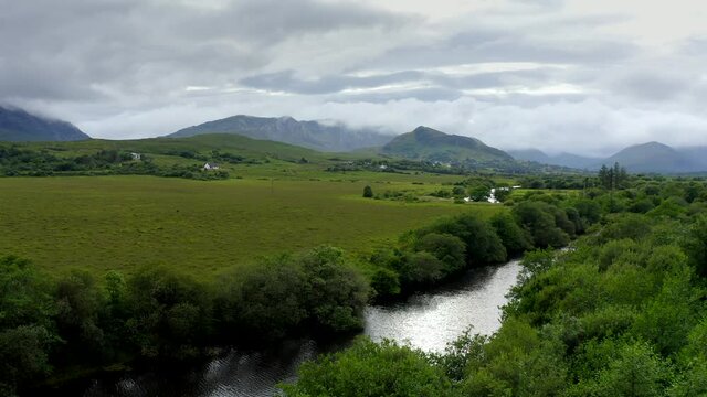 Bealanabrack River, Maum, Connemara, County Galway, Ireland, July 2021. Drone follows the course of the river west over grassy marshland while slowly ascending and revealing Maumturk Mountains.