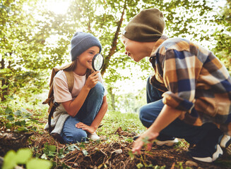 Smiling girl looking through magnifying glass at boy while squatting together on ground in forest