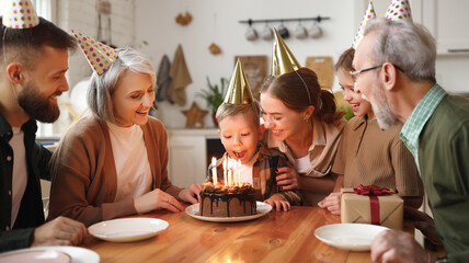 Happy little boy celebrating birthday with family at home, looking at cake with lit candles