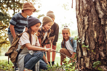 Fototapeta premium Happy school children boys and girls with backpacks examining tree bark in forest