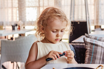 Cute little girl in beige clothes  sits at the table indoors and plays absorbedly with something in her hands,  white table and curtains in the background. Smart activity concept