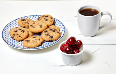 White cup with coffee, traditional cookies and sweet cherries in a bowl on a white wooden background, closeup. Tasty breakfast, morning routine concept