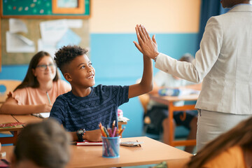 Happy black elementary student and his teacher greet with high five gesture during class at school.