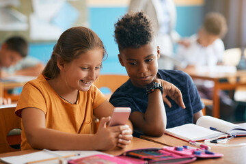 Happy classmates use cell phone in classroom at elementary school.