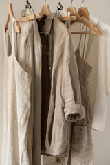 Minimalist women's wardrobe: neutral beige colour washed linen dress or sundress, shirt, pants on hanger against white wall. Aesthetic modern trendy fashion concept