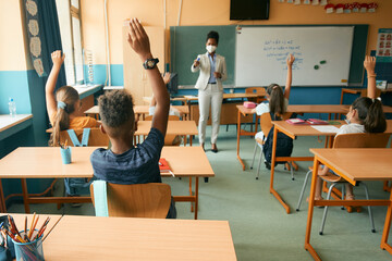 Rear view of elementary students raise their hands to answer teacher's question while having class at school during coronavirus pandemic.