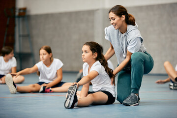 Sports teacher assists her student with stretching exercise during physical education class at school gym.