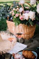 Beautiful picnic outside: wicker basket full of summer flowers, rose wine, coffee to go, croissants and other pastry, hat, sunglasses, book, cherries on the green grass - 451930751