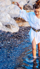 Beautiful fashion caucasian girl with wavy hair, wearing white dress and walking in the sea water, rocky coast. Concept of fashion and travel photo session. Hight quality photo