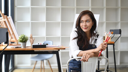 Beautiful Asian woman artist holding paintbrush and sitting at her workshop.