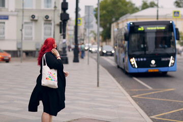 European plus size woman waiting transport at bus stop. Young red pink haired body positive girl.