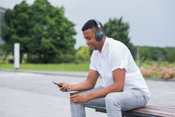 Portrait of a young and happy African American man with headphones. A man sitting on a bench and listening to music, holding a smartphone and a mug of coffee