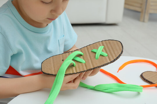 Little Boy Tying Shoe Lace Using Training Cardboard Template At White Table Indoors