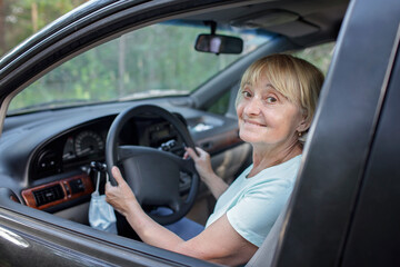 Fototapeta na wymiar Happy senior woman driving a car, medical mask hanging on the steering wheel, solo traveler, independent and active retirement