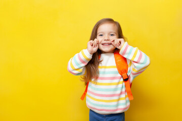 Portrait of a cute little schoolgirl with a backpack in a jumper on a yellow colored background. Back to school. Academic year, September. Training, development, education. A place for text.