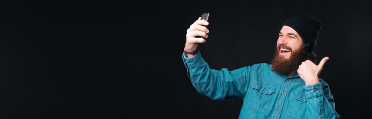 Bearded male hipster is making a selfie with one thumb up.