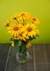 a bouquet of yellow heliopsis flowers stands in a glass vase on a wooden table with a green wall. side view