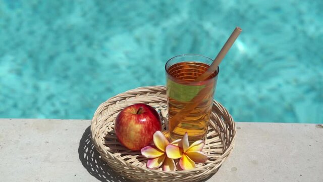 Video footage of glass with apple juice, bamboo straw, red apple, tropical flower frangipani and bubbling blue swimming pool on background.